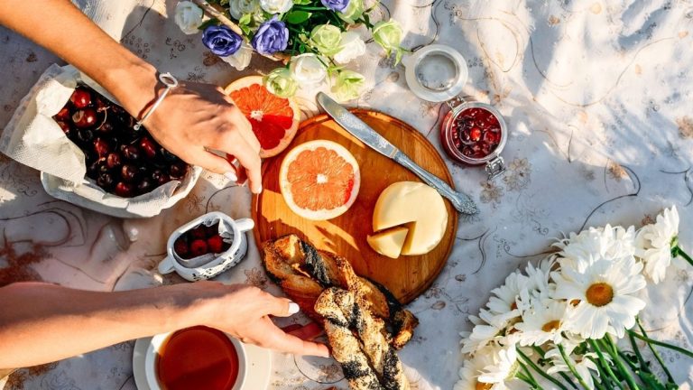 Picnic essentials for adults elegantly laid out on a blanket with fruits, cheese, bread and flowers.