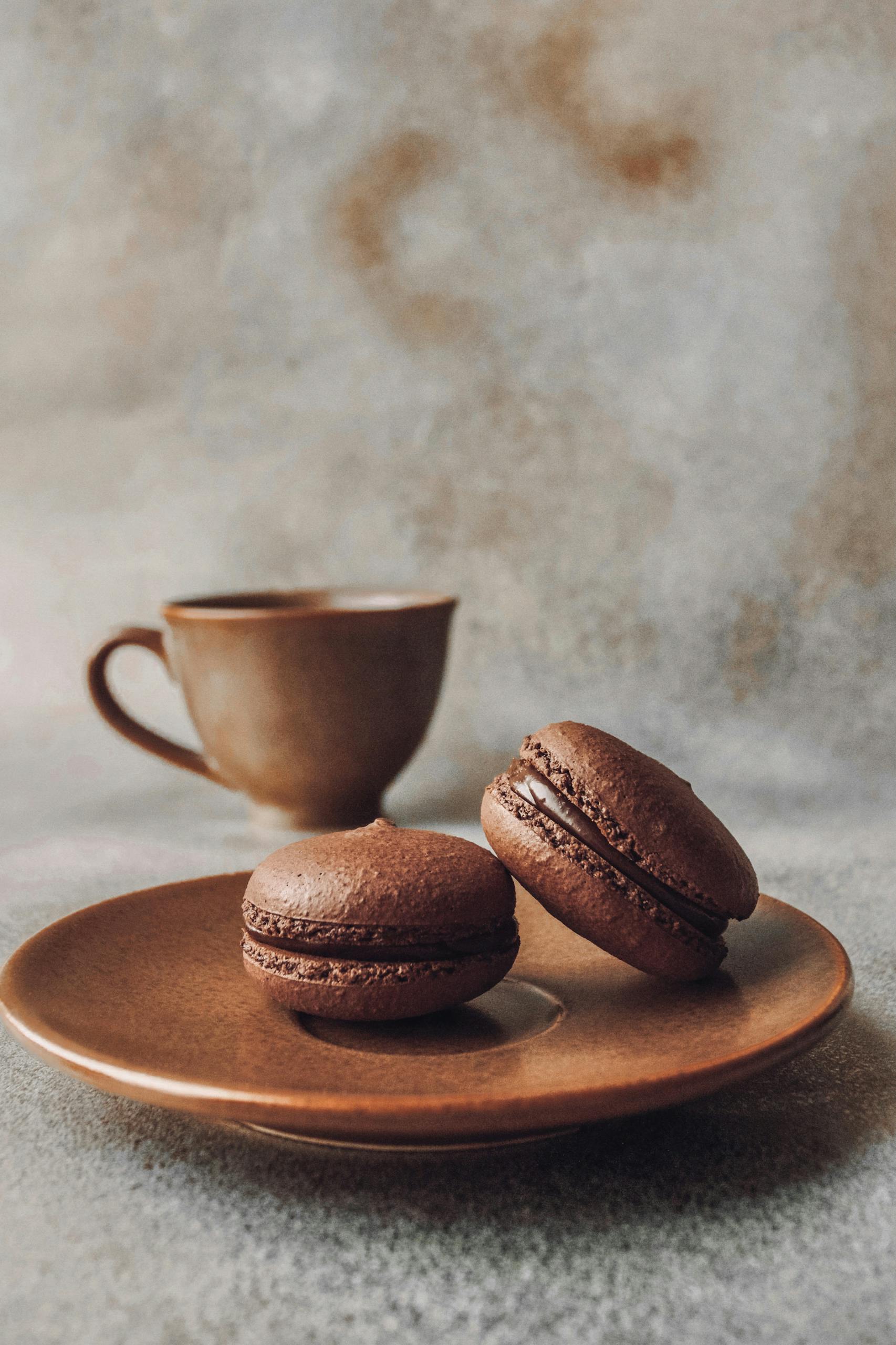 Delicious chocolate macarons on a brown ceramic saucer with a coffee cup in a rustic setting.