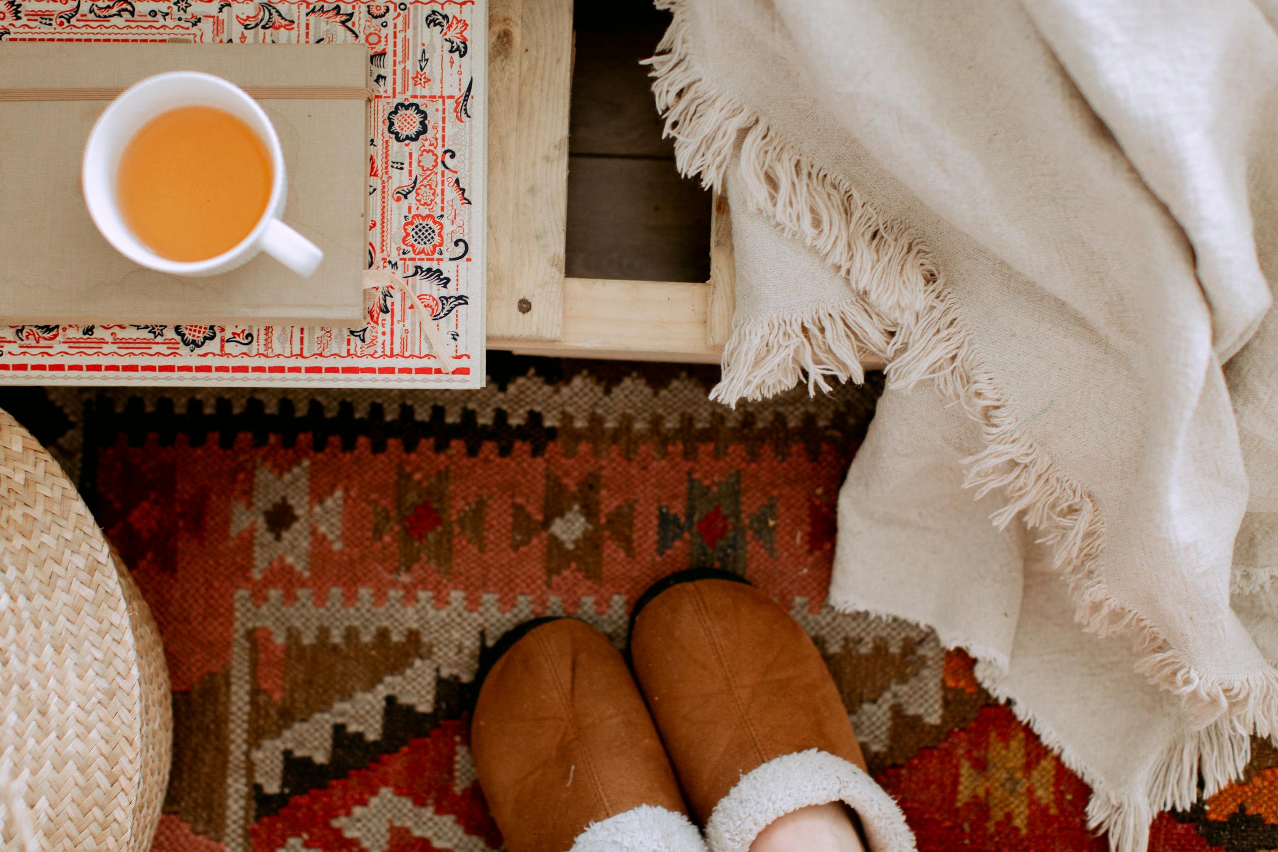 From above of cozy bedroom interior with white plaid, brown warm slippers on carpet, wicker basket and cup of tea on tray for cozy fall outfits lazy days at home.