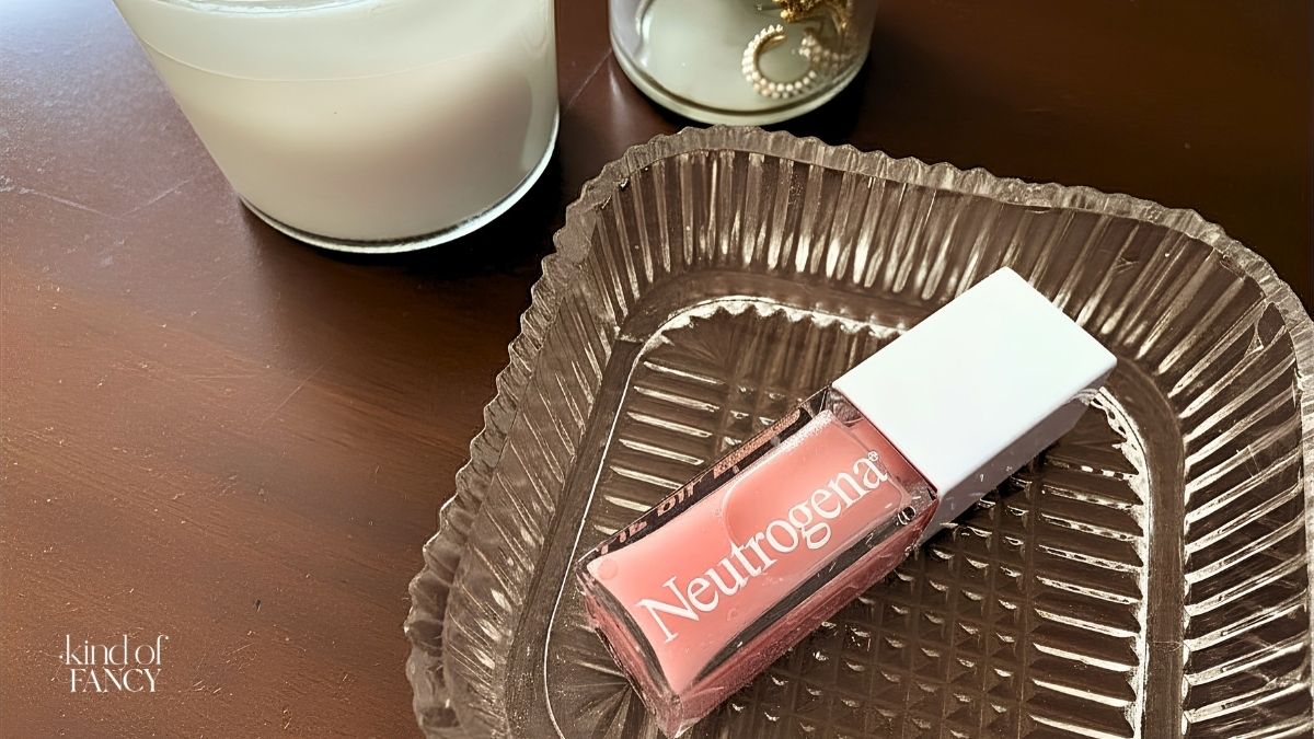 Wooden nightstand table with glass tray holding Neutrogena Hydro Boost lip oil in light pink next to a white candle and pearl huggie earrings.