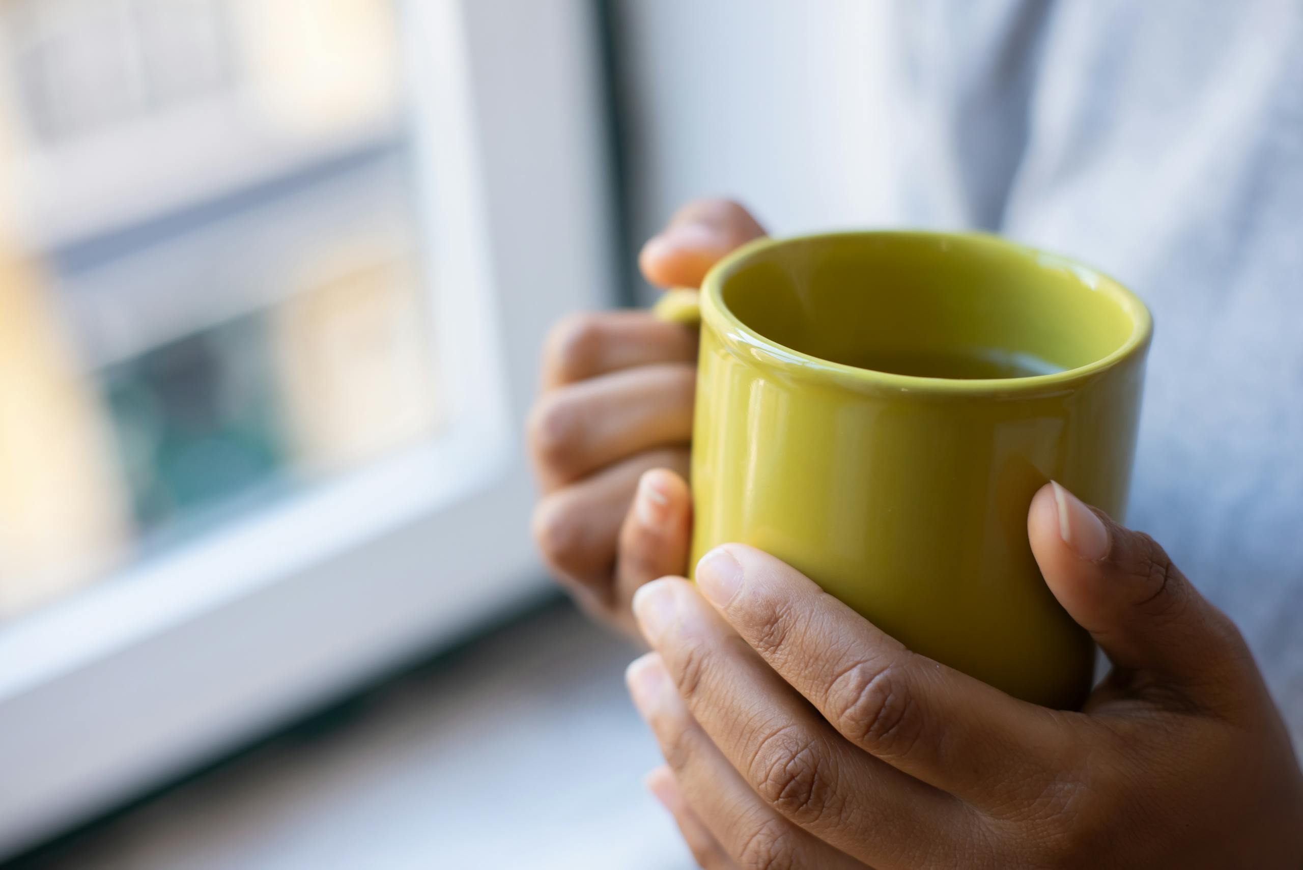 Close-up of pretty hands holding a yellow mug near a window, creating a warm and cozy vibe.